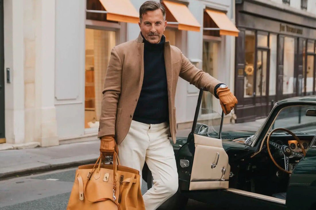 Stylish man in a camel coat and white pants holding a leather bag while getting out of a classic car on a city street.