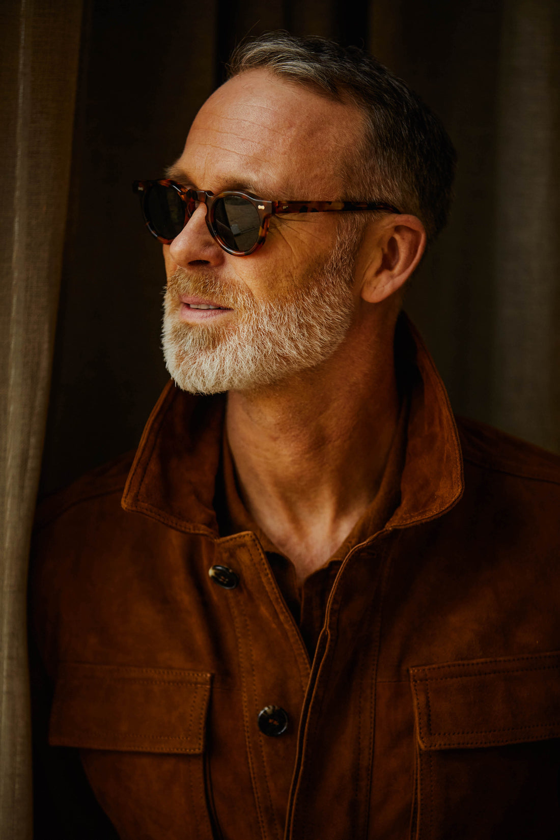 Mature man in sunglasses and brown jacket looking away thoughtfully.