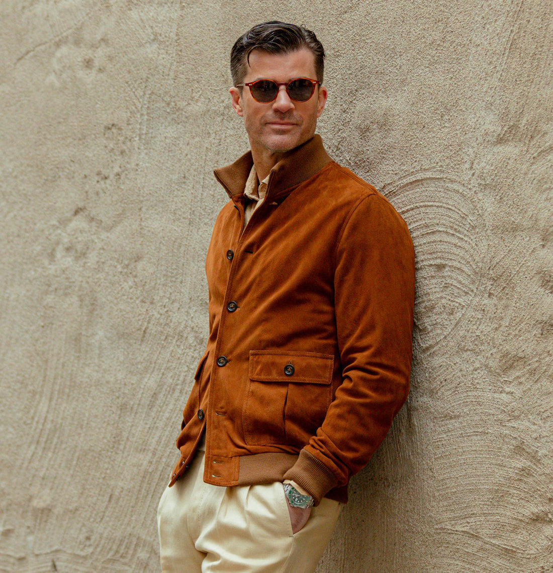 A stylish man in sunglasses, a brown suede jacket, and cream trousers leans against a textured wall.