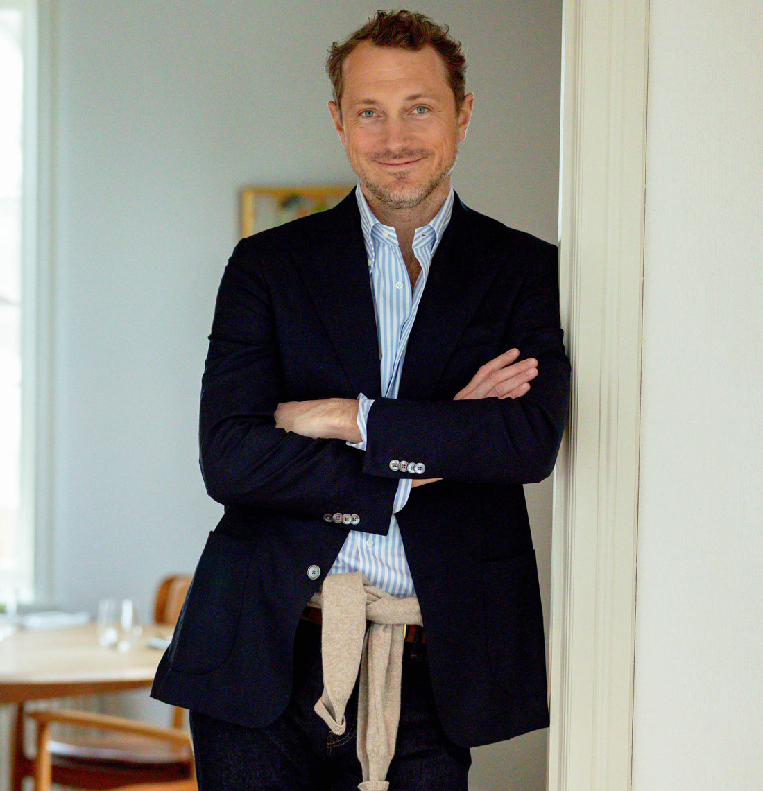 A man in a blazer and striped shirt, arms crossed, smiling and leaning against a doorway in a lightly furnished room.