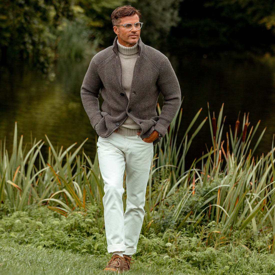 A man in a brown jacket and white pants standing by a pond with greenery in the background.