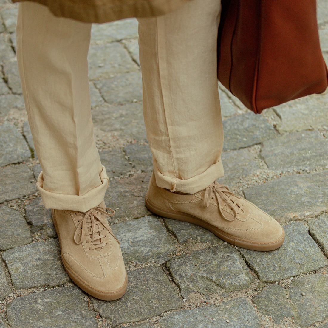 A person wearing beige trousers and beige lace-up shoes stands on a cobblestone path, holding a brown leather bag.