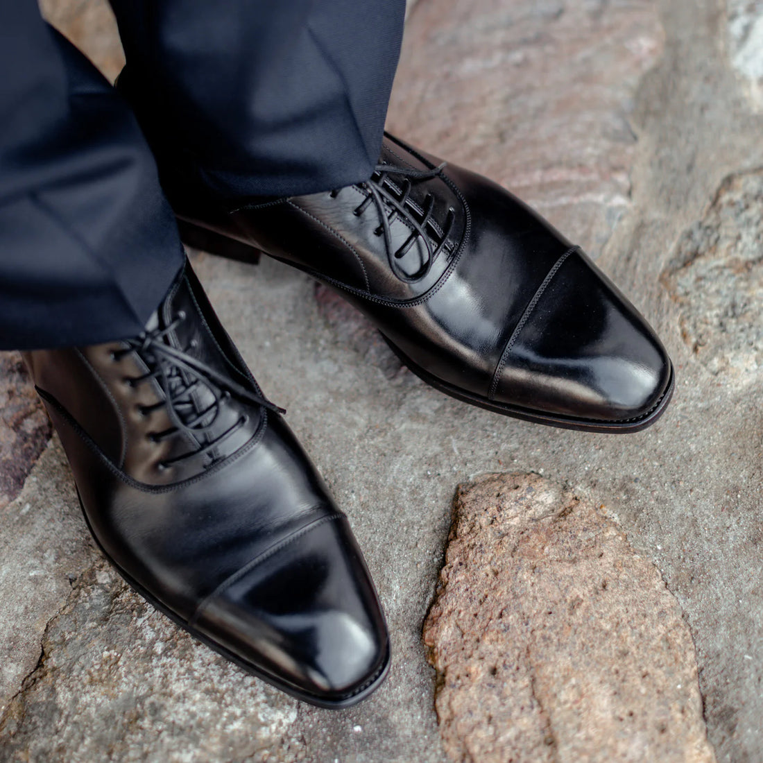 Close-up of a person's feet wearing polished black dress shoes, standing on a stone-paved surface.