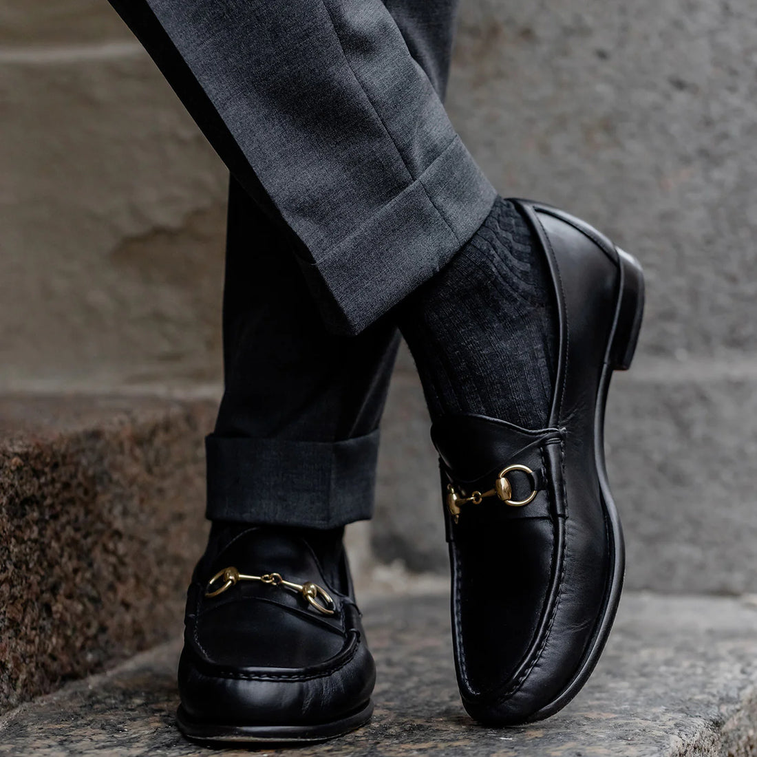 Close-up of a person's feet wearing black loafers with gold accents and grey trousers on a stone step.