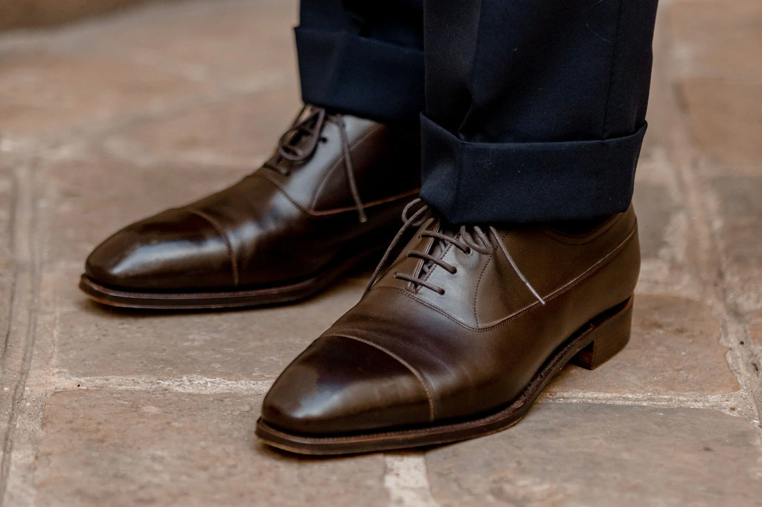 A close-up of a pair of polished brown leather shoes worn with dark trousers.
