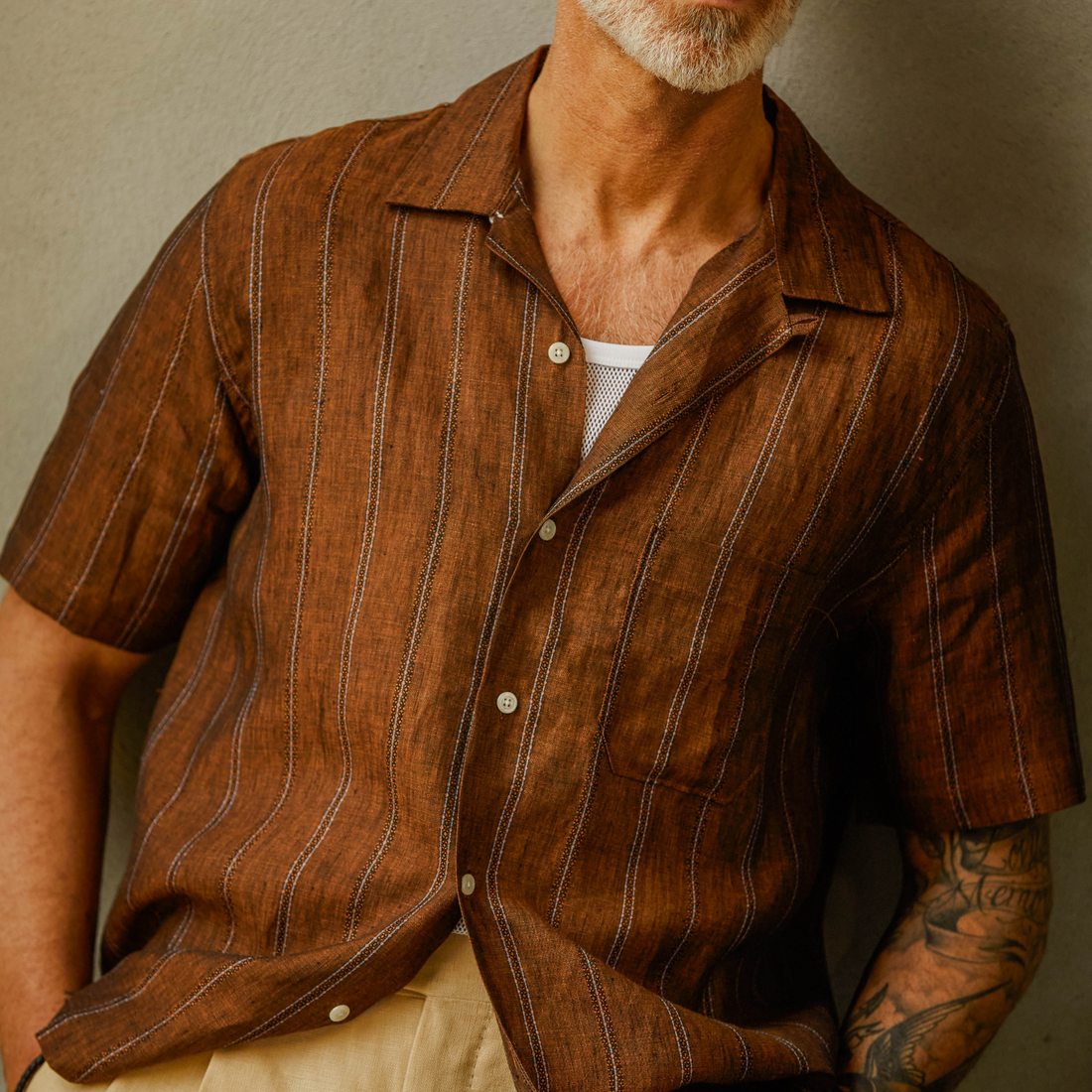 An older man with tattoos on his arms wearing a striped brown shirt and beige pants, cropped at the mid-chest.