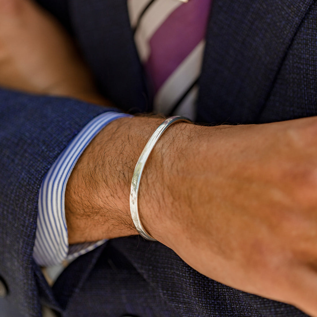 Close-up of a man's wrist wearing a silver bracelet, with a glimpse of his blue-striped shirt cuff and part of a navy suit.
