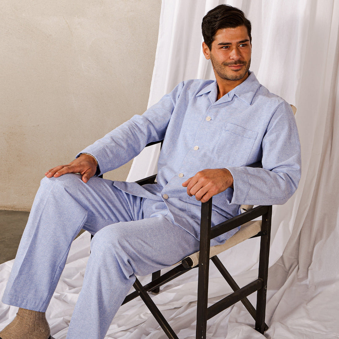 A man in a light blue jumpsuit sits on a black director's chair, smiling slightly, against a draped white background.