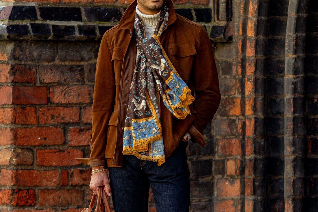 A man wearing a brown jacket, decorative scarf, and jeans standing against a brick wall.
