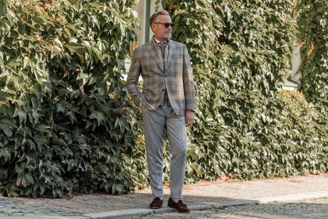 Man in a checkered blazer and trousers standing on a cobblestone path beside green foliage.