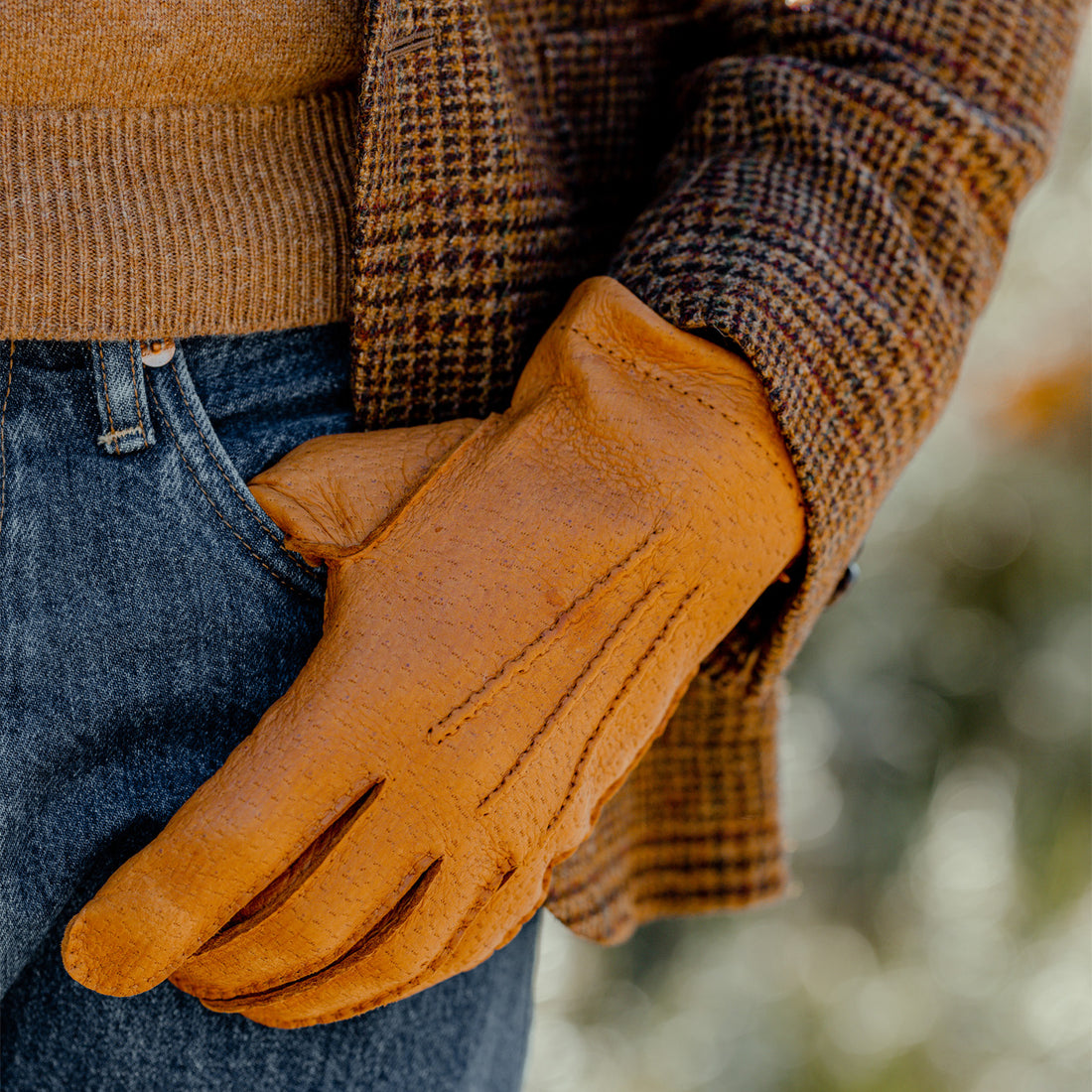 A person wearing a brown sweater and denim jeans tucking a pair of leather gloves into their pocket.