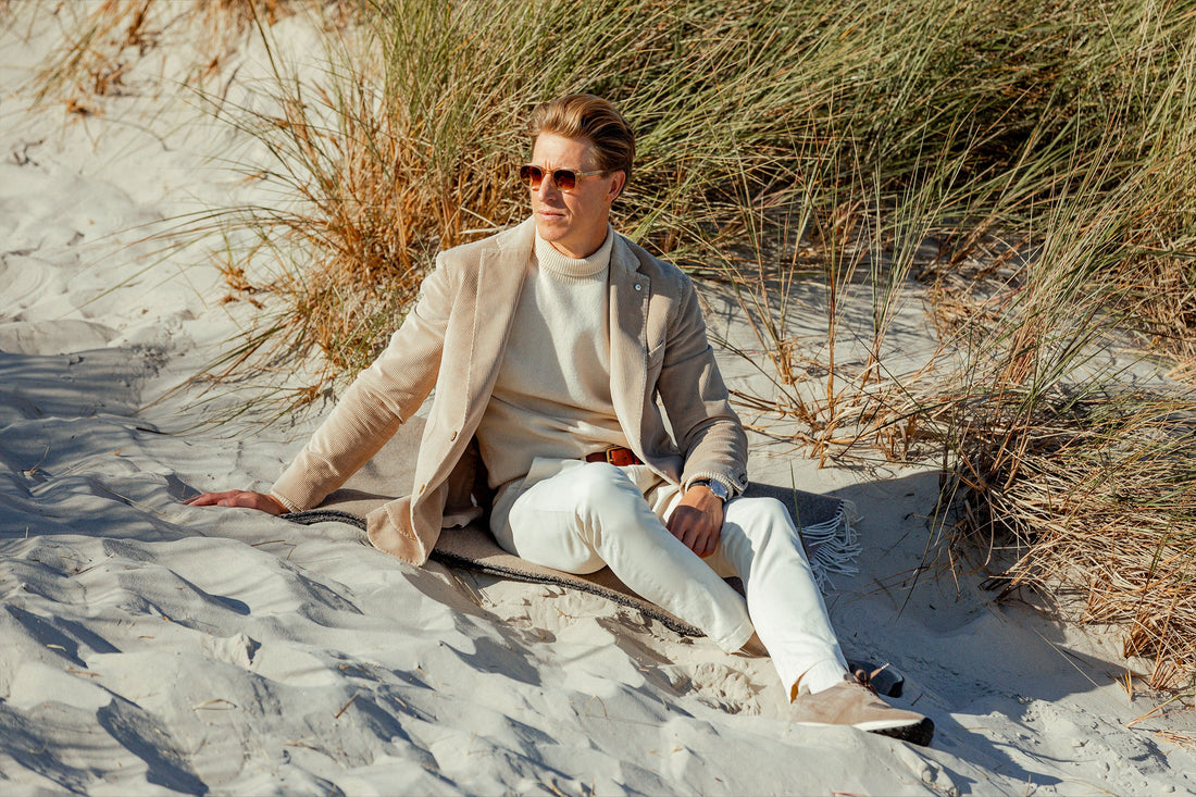 A man in smart-casual attire sitting relaxed on a sandy beach with dune grass around him.