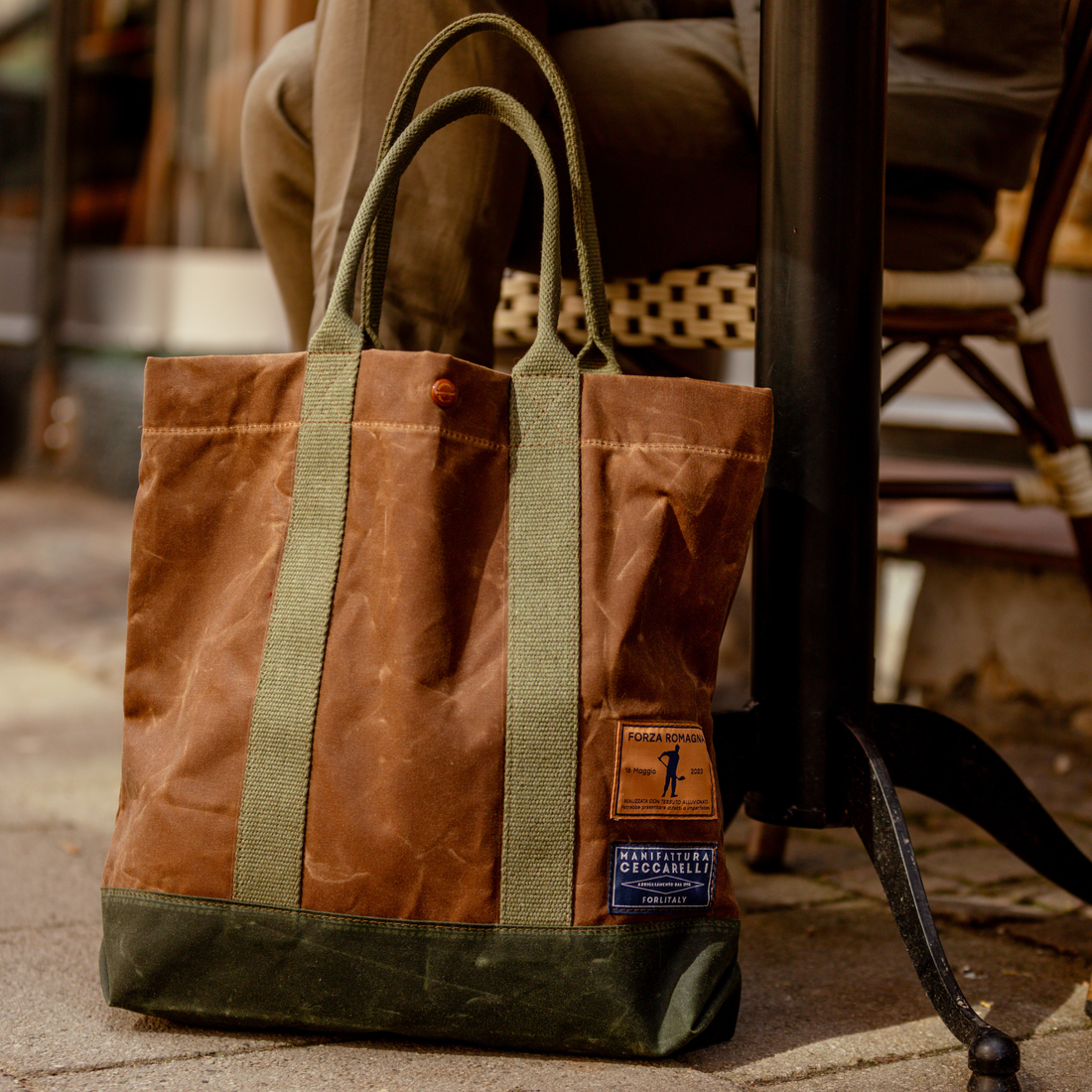 A worn leather and canvas tote bag with visible branding stands next to a seated person, placed on a sidewalk.