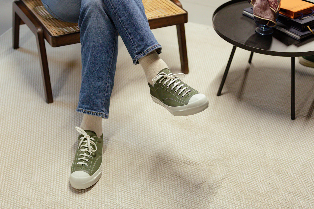 A person wearing green sneakers and blue jeans with their feet elevated above a patterned carpet.