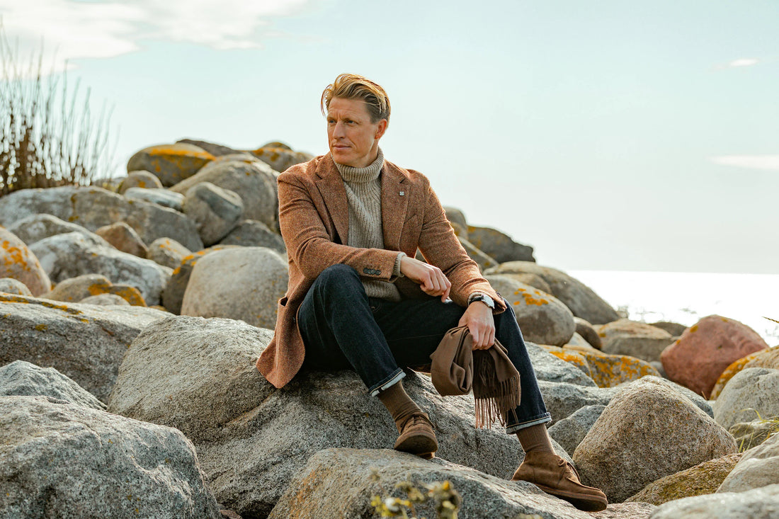 A man seated on a rocky shore dressed in autumn attire, looking pensively into the distance.