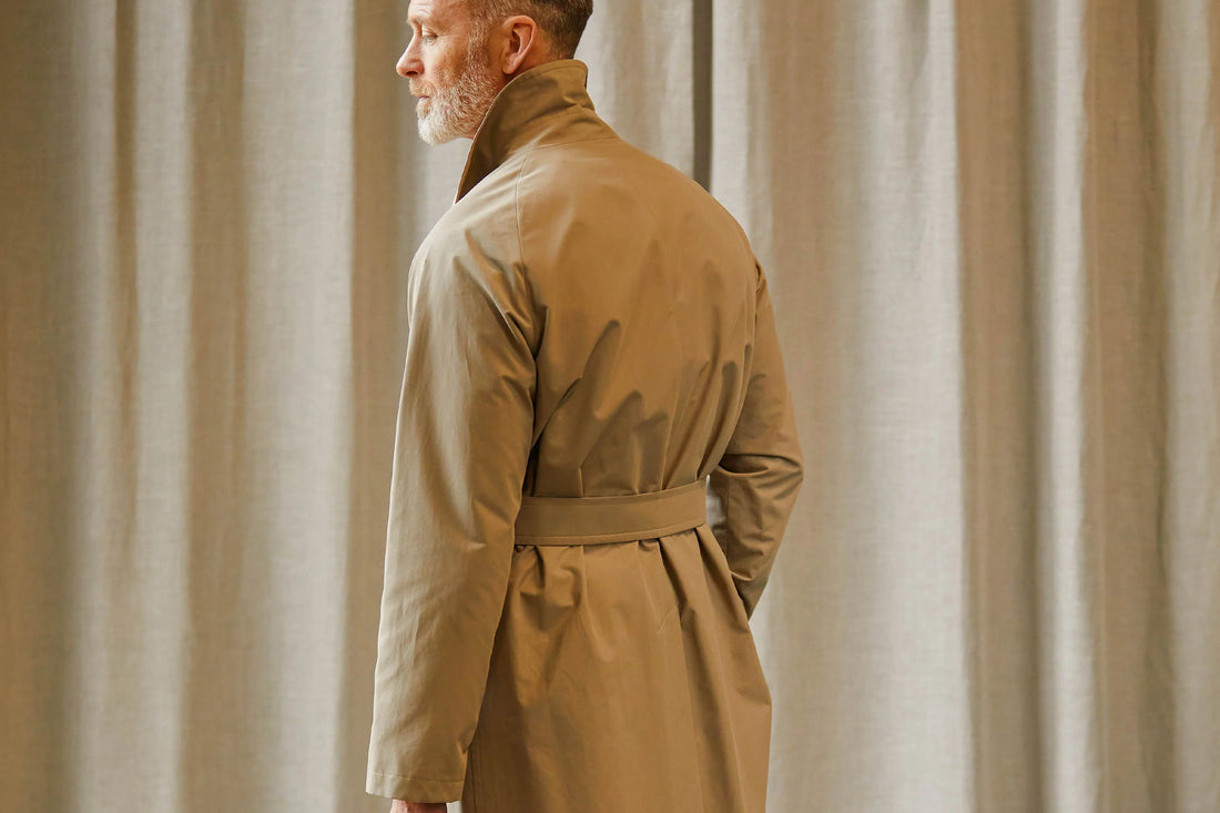 Man in a beige trench coat standing sideways with a neutral backdrop.