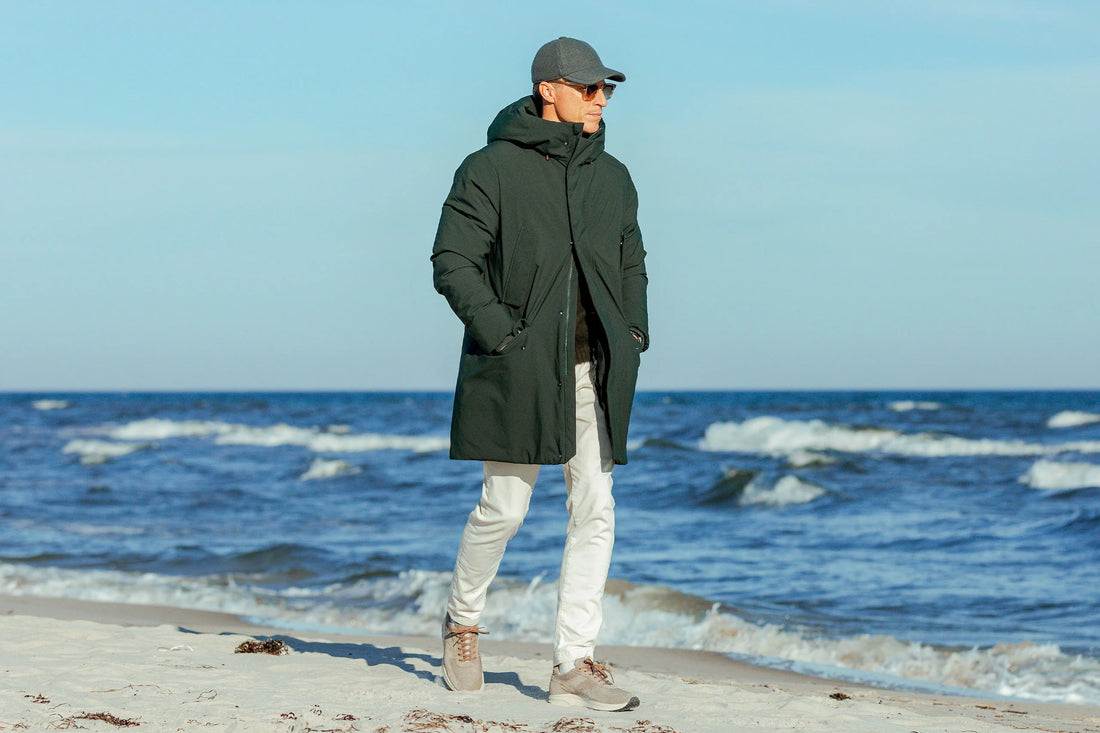 A person in a winter coat and sunglasses walking along the shore with waves in the background.