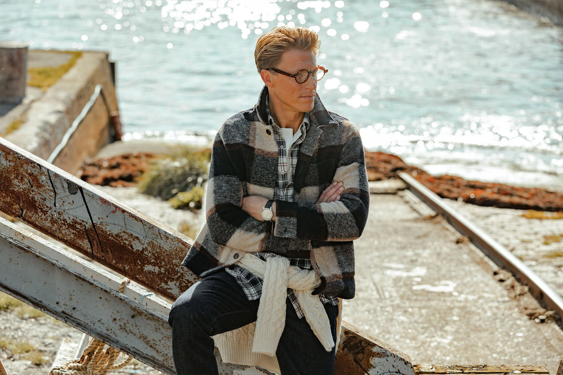 Man sitting on a metal railing by the waterfront, wearing a checkered jacket and sunglasses.
