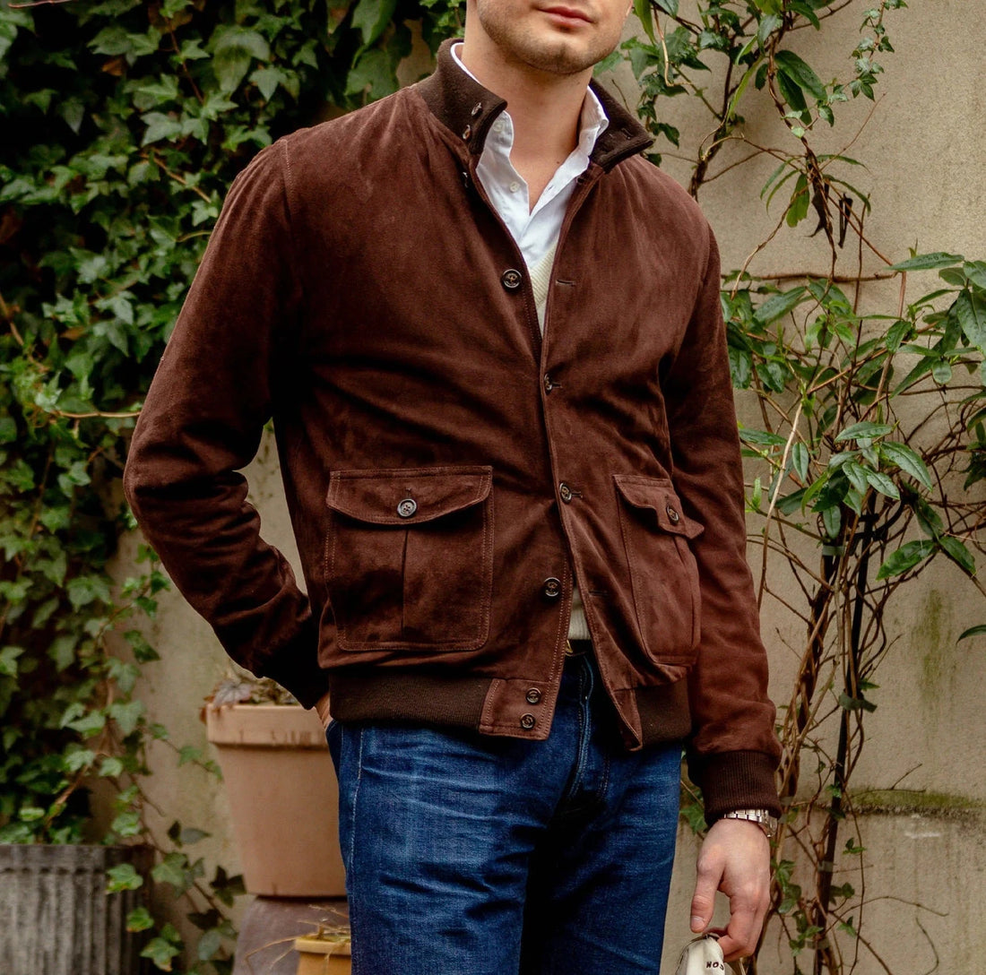 Man wearing a brown suede jacket and blue jeans standing by a wall with foliage.