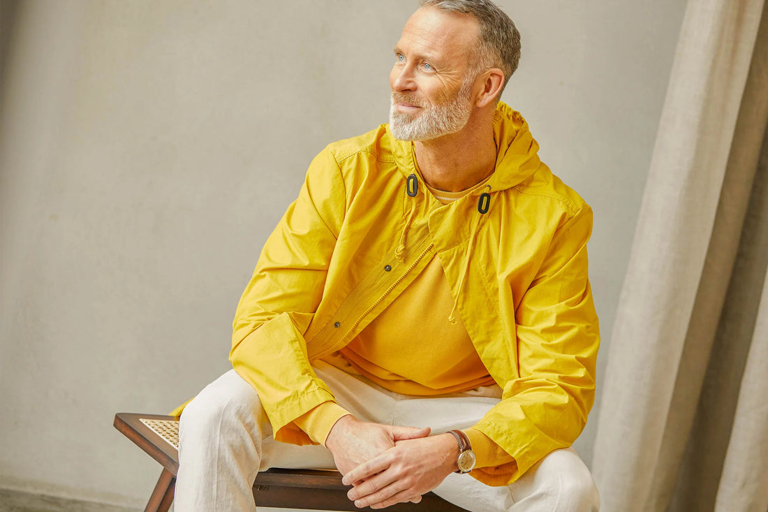 A smiling middle-aged man in a yellow jacket sitting on a stool, looking to the side.