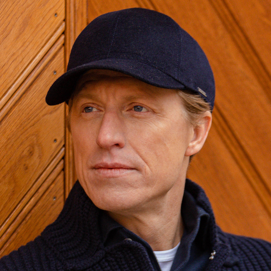 A man wearing a cap looks off into the distance, with a wooden door in the background.
