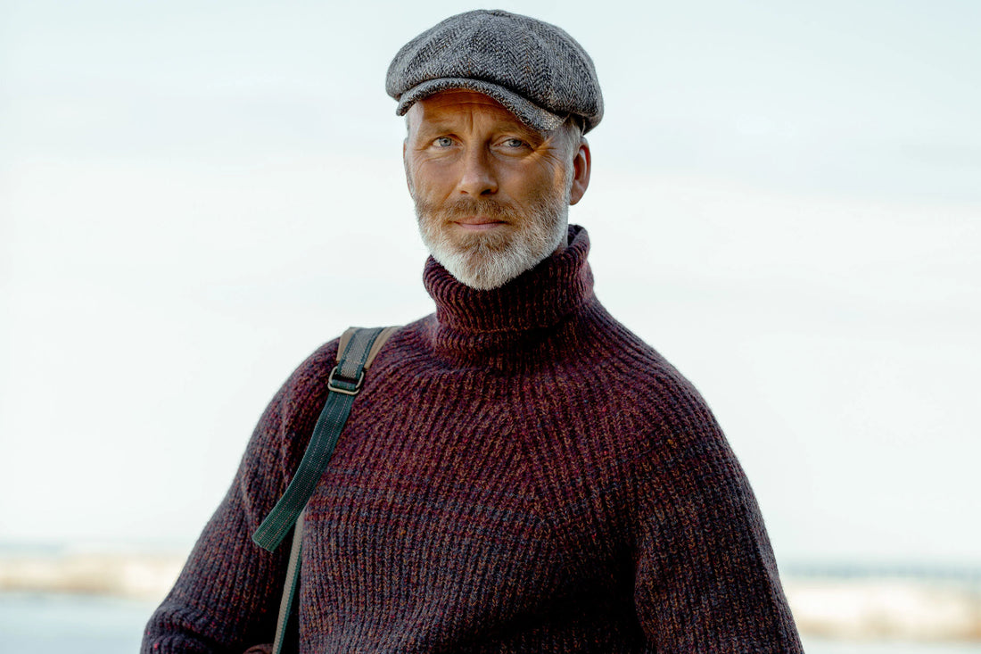 A portrait of a bearded man wearing a turtleneck sweater and flat cap, with a shoulder bag and looking at the camera.