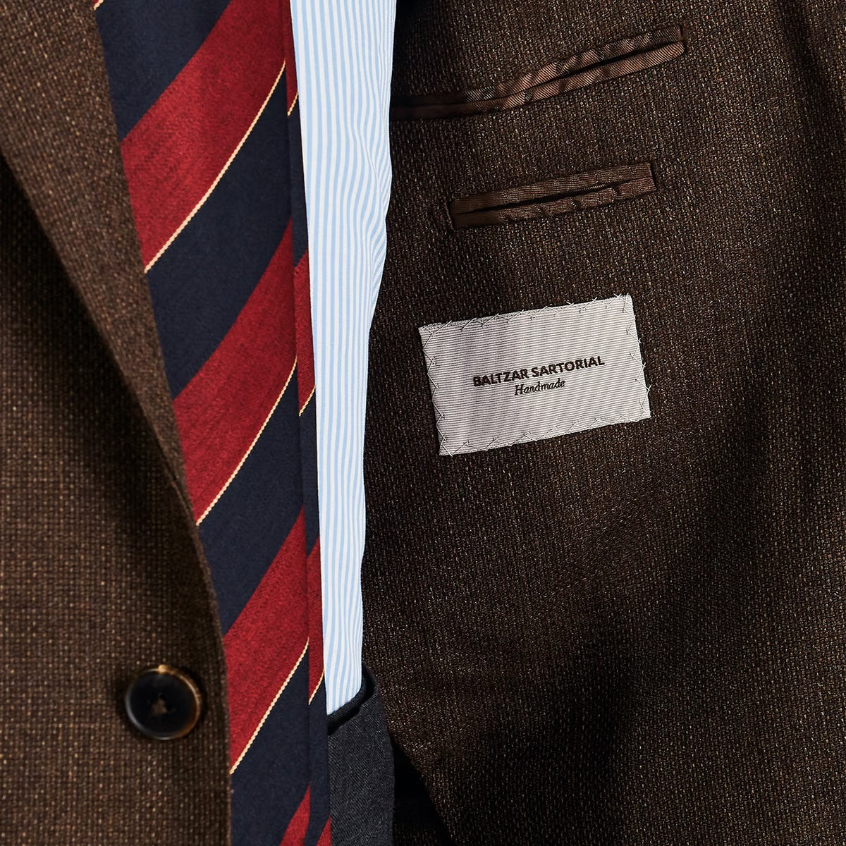 Close-up of a brown tweed jacket with a "Baltzar Sartorial Handmade" label, partially visible striped shirt, and a red and blue striped tie featuring Baltzar Sartorial tailored clothing.