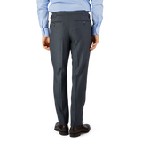 A man wearing a blue shirt and Grey Super 100's Wool Flat Front Suit Trousers by Baltzar Sartorial, standing on a black surface against a blue and gray background.