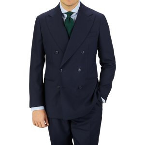Man wearing a Baltzar Sartorial Navy Blue Super 100s Wool DB Suit Jacket with a green sweater and dark blue tie, standing against a light gray background.