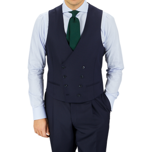 Man in a tailored Baltzar Sartorial Navy Super 100s Wool DB Waistcoat, white shirt, dark tie, and matching trousers, standing against a gray background.