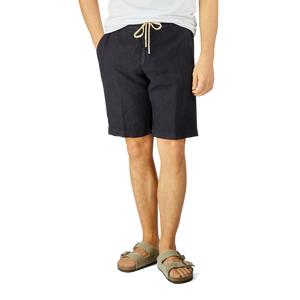 Man wearing Berwich navy blue washed linen drawstring shorts and gray sandals stands against a light background, focusing on his lower torso and legs.