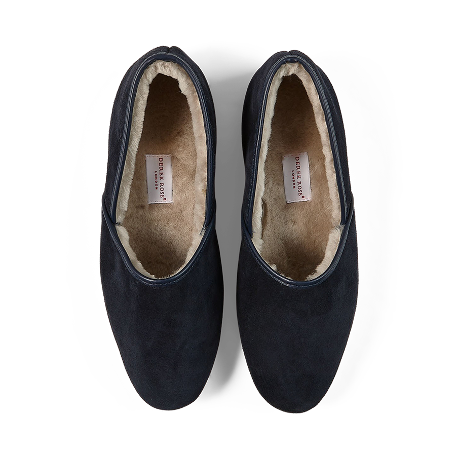 A pair of handmade Navy Suede Sheepskin Closed-Back Slippers from Derek Rose against a black background.