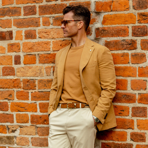 A man in a L.B.M. 1911 Dark Beige Washed Cotton Linen Blazer and sunglasses leaning against a brick wall.