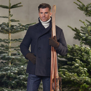 A man in a Navy Blue Heavy Wool Casentino Peacoat by Manifattura Ceccarelli holding skis in front of a Christmas tree.
