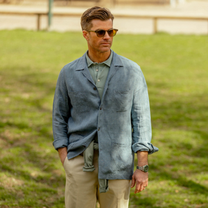 Man in sunglasses and a Mazzarelli Indigo Blue Organic Linen Four Pocket Overshirt standing in a park, looking to the side with a thoughtful expression.