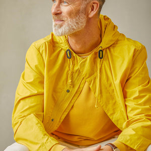 Mature man with a beard wearing a water-resistant Yellow Cotton Ripstop Stanedge Jacket by Universal Works, sitting and looking to the side.