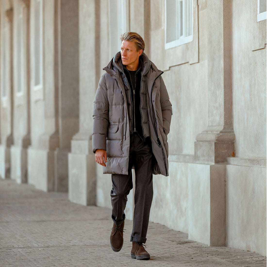 A man walks down a corridor wearing a long grey coat, dark trousers, and brown shoes. The background features columns and windows on a building.