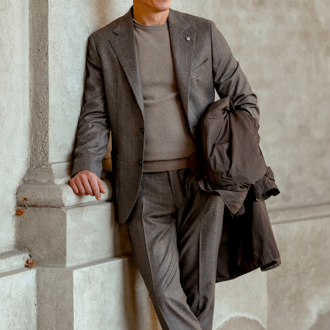 A person in a gray suit leans against a wall, holding a brown coat. The background consists of a concrete structure.