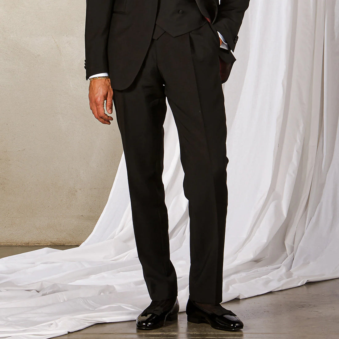 A person in a black suit and shiny shoes stands beside a white draped fabric.