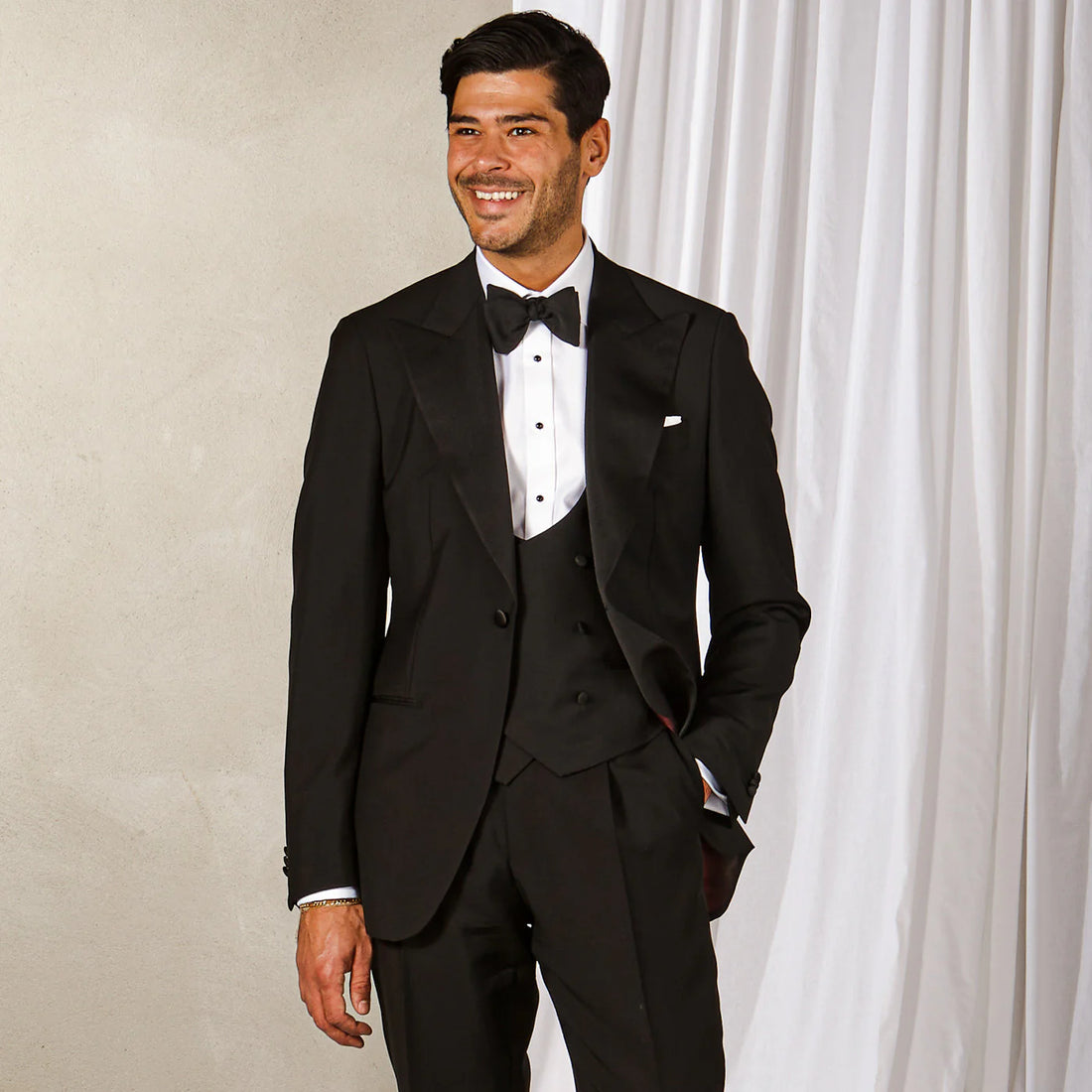 Smiling man in a black tuxedo with a bow tie, standing by a curtain.