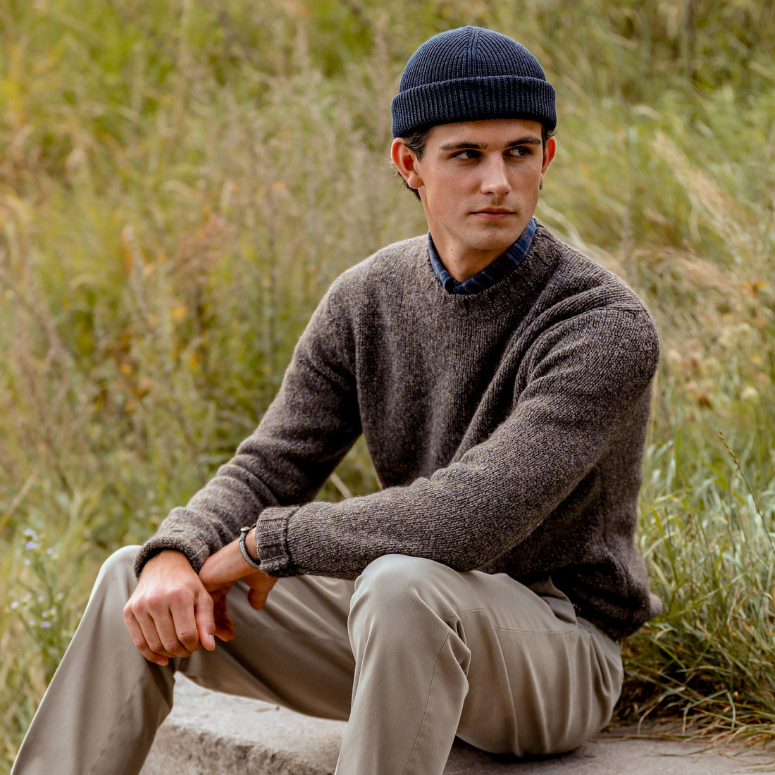 Man seated in a chair wearing a white sweater and gray pants, with his hand on his knee showcasing a watch.