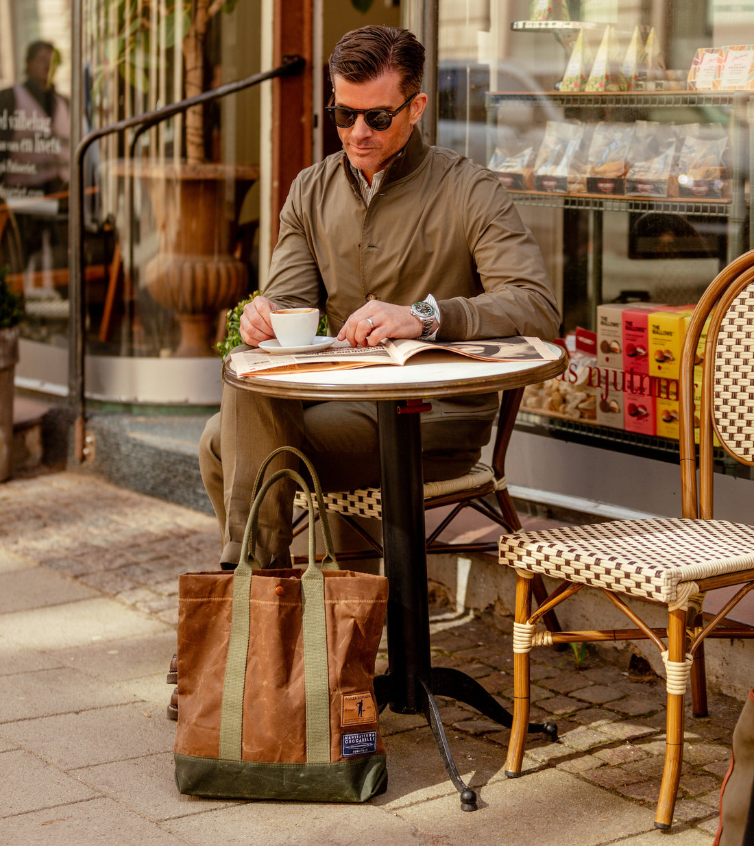 Man in sunglasses sitting at a café table, sipping coffee and reading a newspaper, with shopping bags at his side.