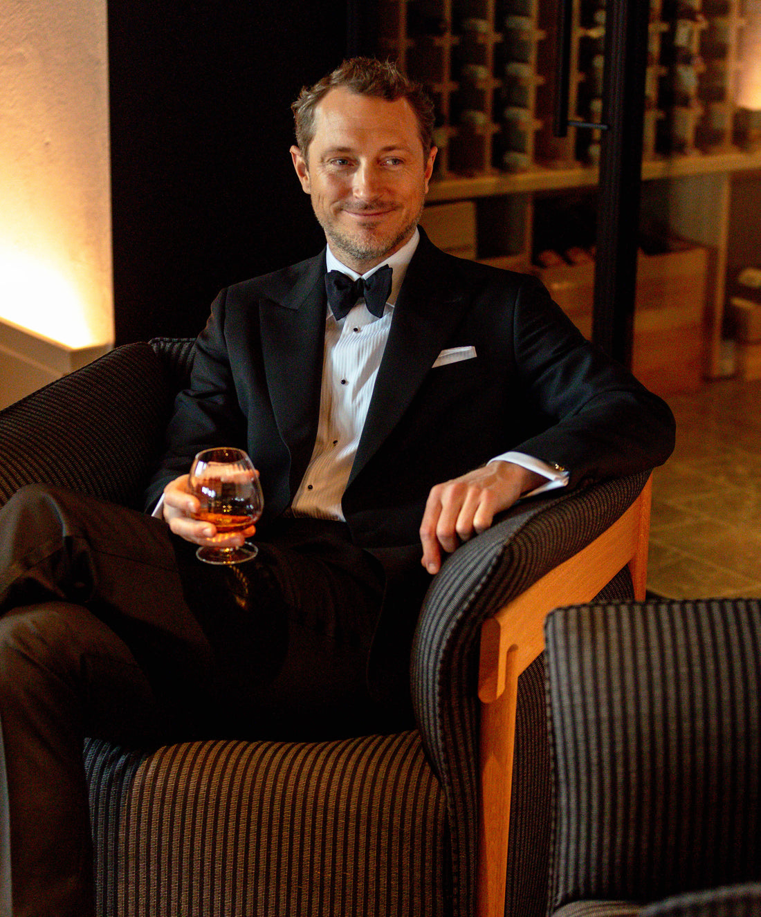 A man in a black tuxedo smiling, sitting in a dark chair with a glass of amber liquid, in a dimly lit room.