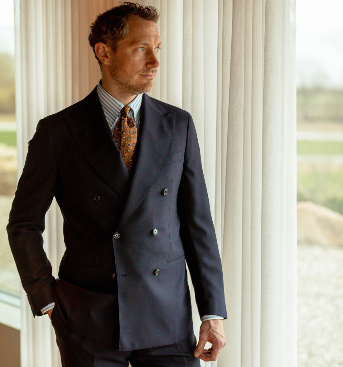 A man in a tailored double-breasted suit stands smiling against a backdrop of a serene coastal landscape.