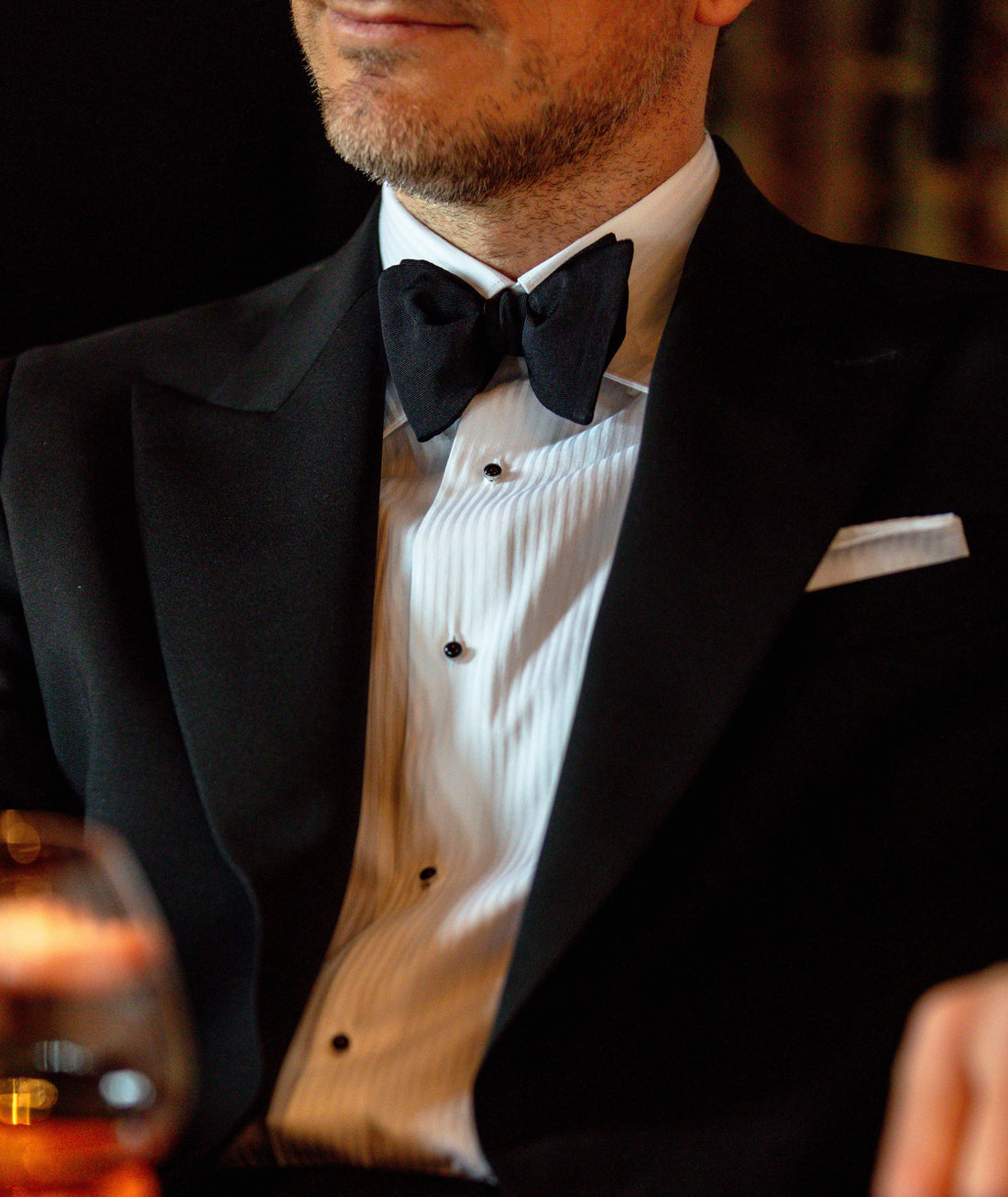 A man in a tuxedo with a bow tie and white shirt, holding a glass of wine, partially cropped to show from his chin down.