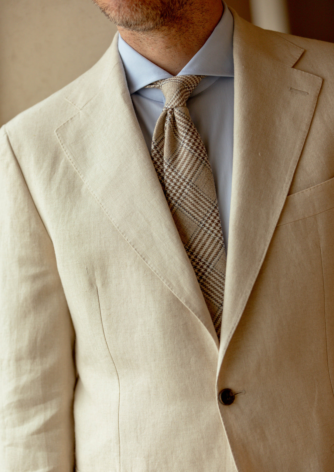 Close-up of a man’s chest wearing a beige suit with a plaid tie and light blue shirt.