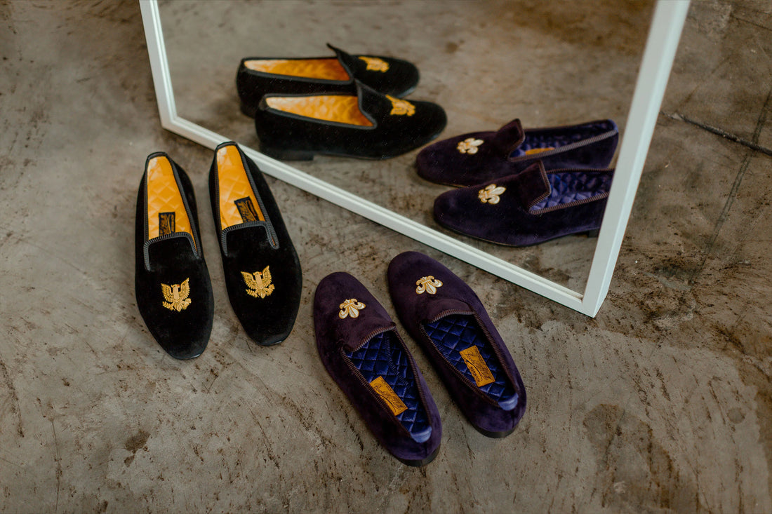 Four pairs of velvet loafers with embroidered designs reflected in a mirror on a concrete floor.