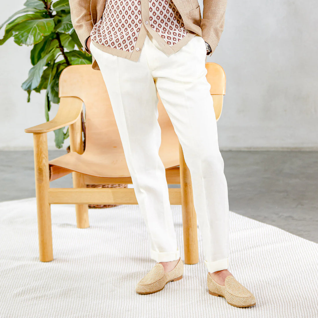 A person stands in white pants and patterned top, with beige loafers, next to a wooden chair in a bright room with a plant.