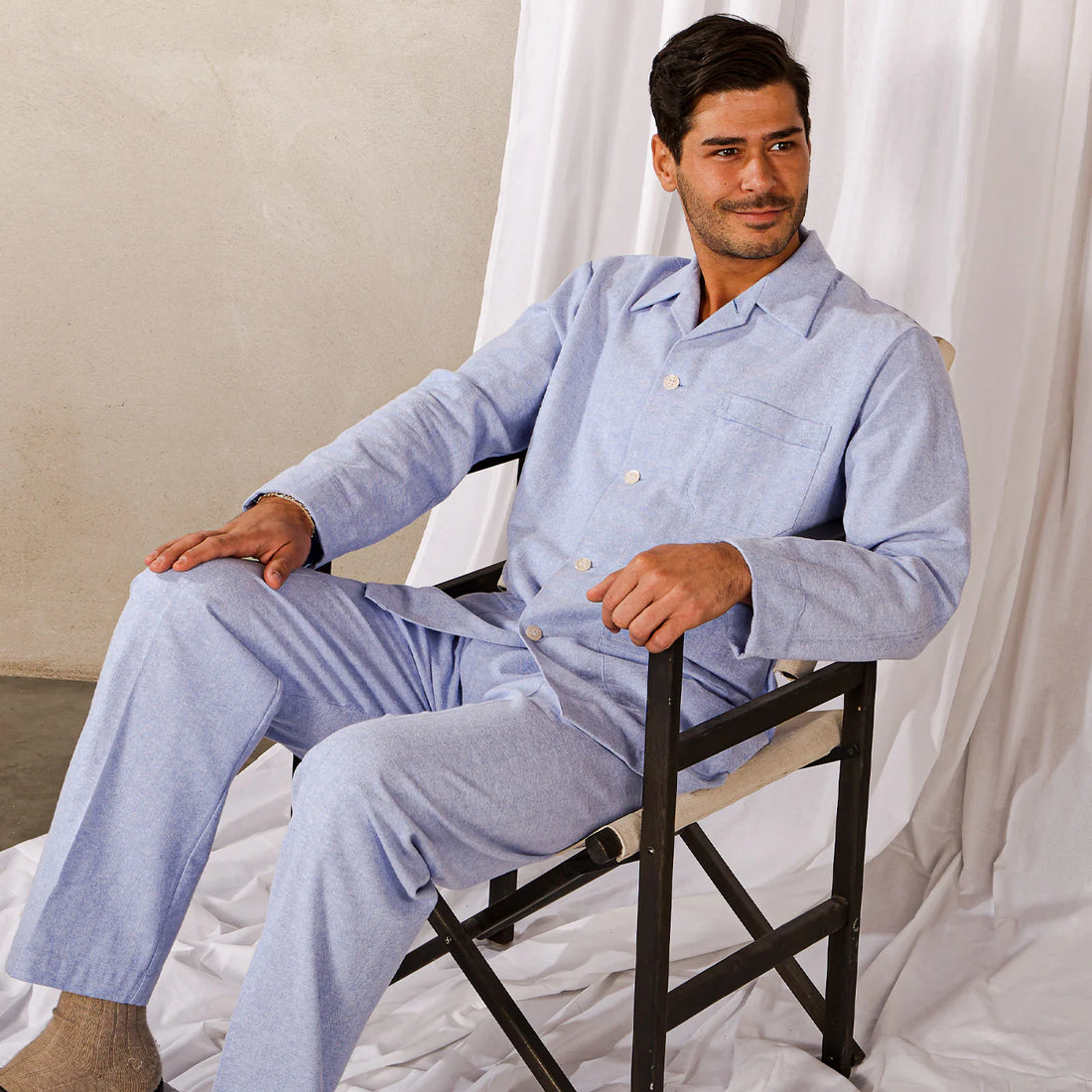 A man in a light blue pajama set sits relaxed on a black folding chair, with a white draped background.