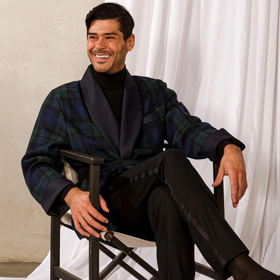 A smiling man in a tartan blazer and black turtleneck sits relaxed in a director's chair against a draped white background.